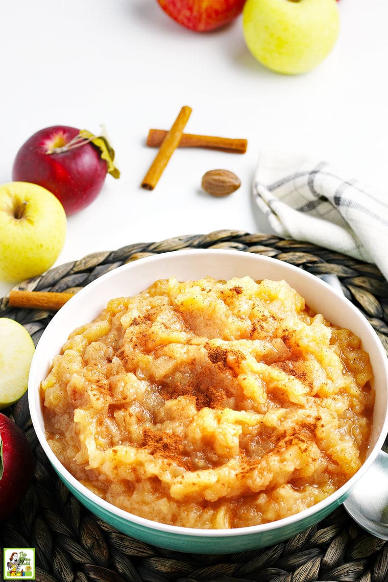 A large bowl of applesauce with a black and white dishtowel, apples, and cinnamon sticks.