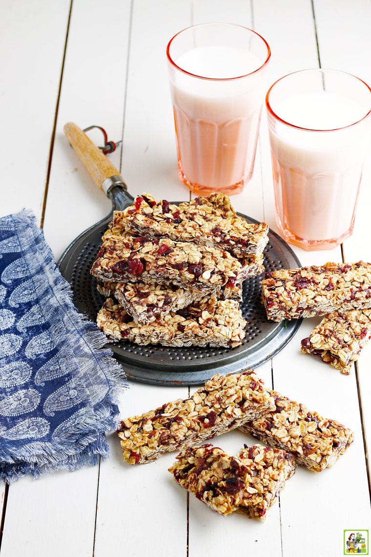 A stack of granola bars with chocolate chips and dried cranberries on a metal trivet with a blue and white napkin and two pink glasses of milk on a white wooden countertop. 