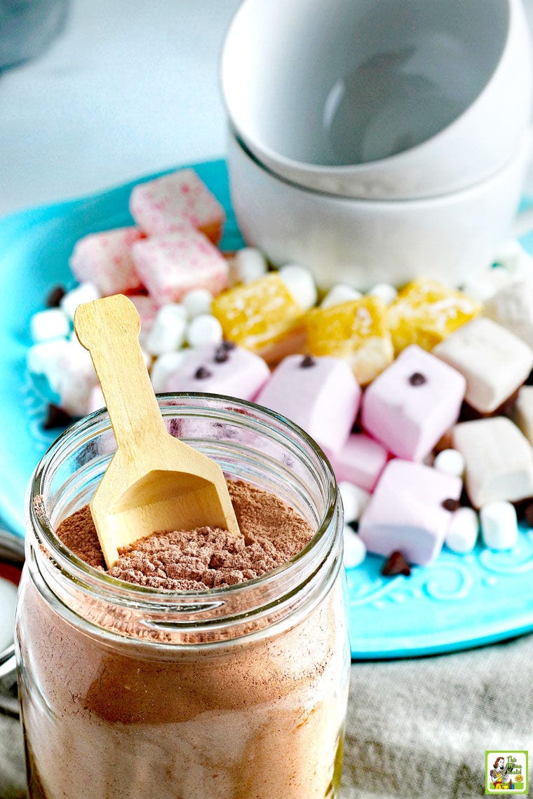 A glass jar of Homemade Hot Chocolate Mix with a wooden scoop. A blue plate of marshmallows, chocolate chips, and white mugs in the background.