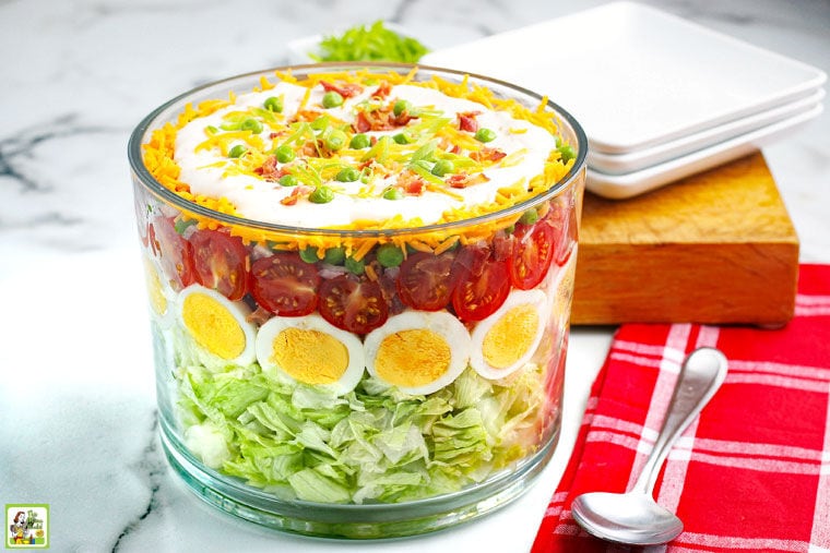 A glass trifle bowl of Seven Layer Salad with serving spoon, red napkin, white plates, and wooden cutting board.