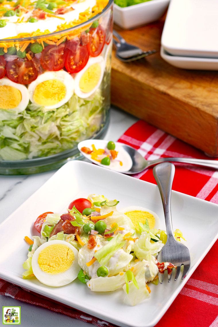 A white plate of Seven Layer Salad with fork, a red napkin, a big glass bowl of layered salad, a wood cutting board, and more white plates.