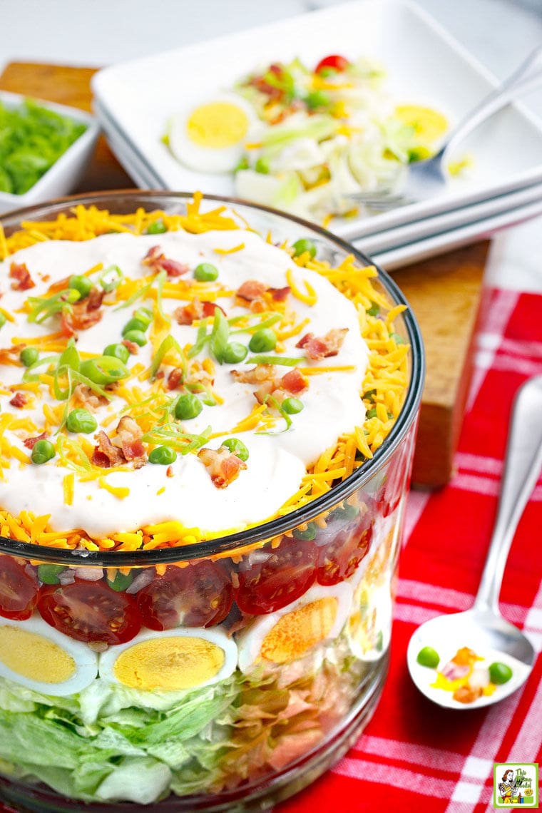 A trifle bowl filled with seven layer salad filled with vegetables, eggs, cheese, and dressing, serving spoon, red napkin, and plates.