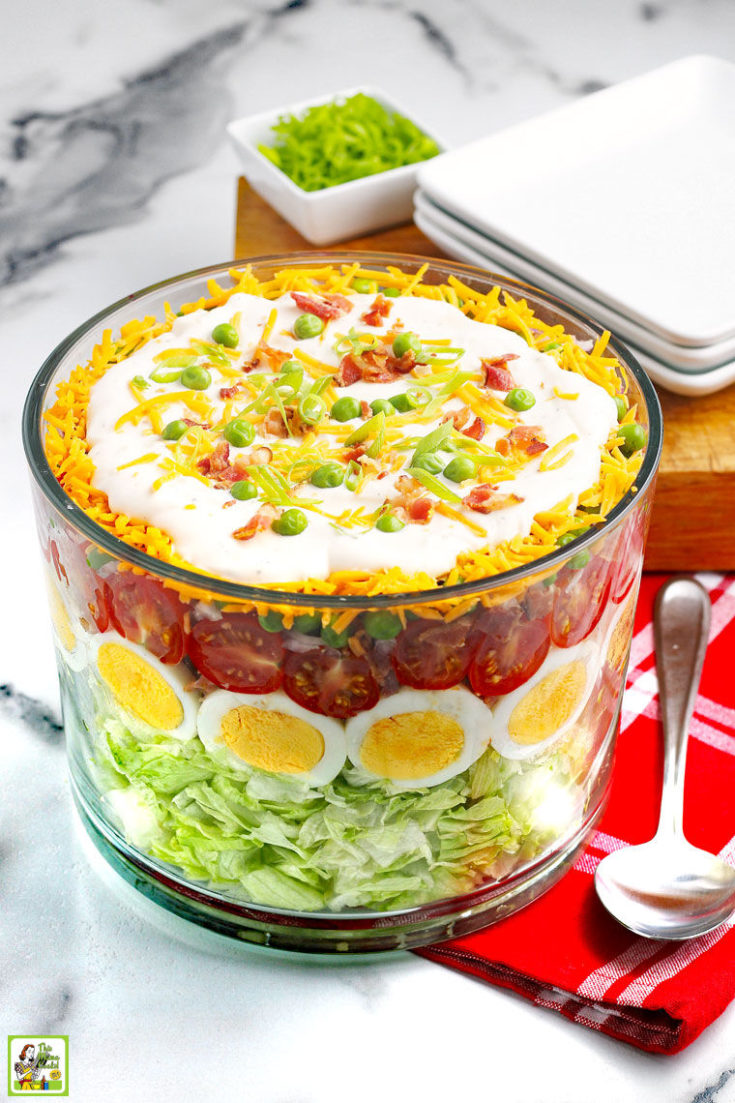 A large glass bowl with a layered salad filled with eggs, tomatoes, peas, cheese, onions, and dressing, plates, serving spoon, and wooden cutting board on a red napkin.
