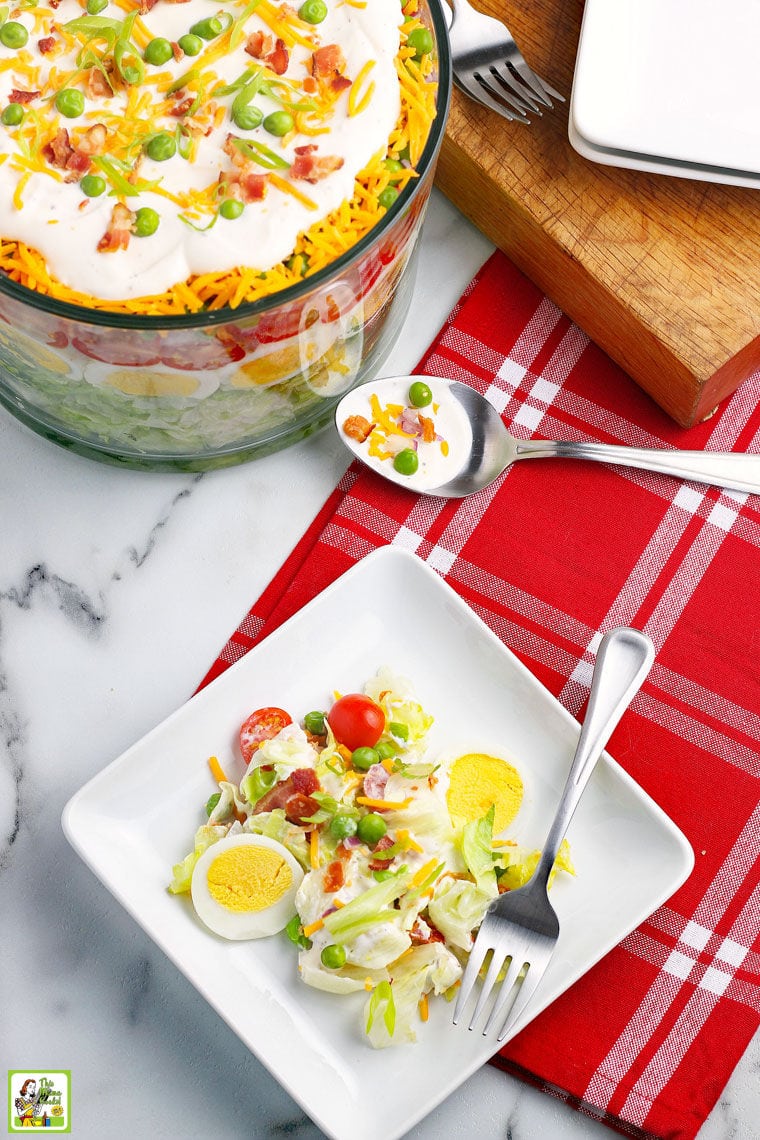 Overhead shot of a plate of salad with fork, red napkin. a large glass bowl of layered salad, a serving spoon, and a wooden cutting board. 