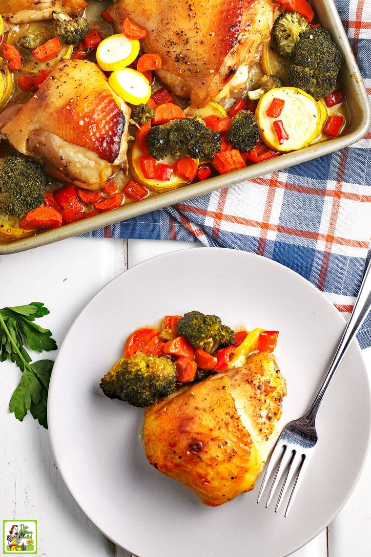 Baked chicken thigh and roasted vegetables on a white plate with a fork, plaid napkin, and a baking sheet with more chicken and roasted vegetables.