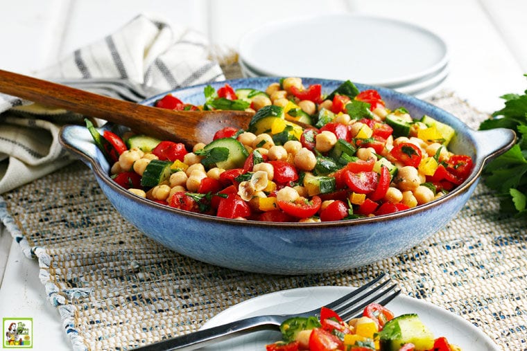 Colorful garbanzo bean, tomato, and cucumber salad in a blue bowl with a wooden serving spoon.