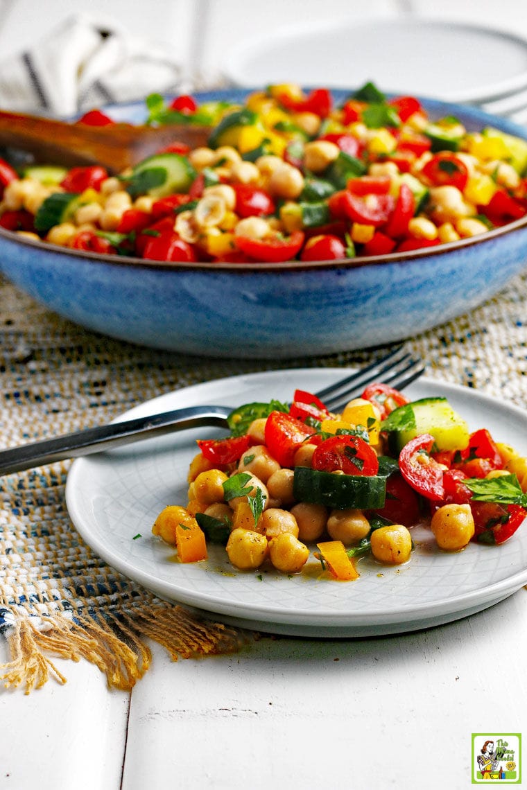 A blue bowl and white plate with fork with garbanzo bean salad on a rustic grey woven placemat with white plates in the background.