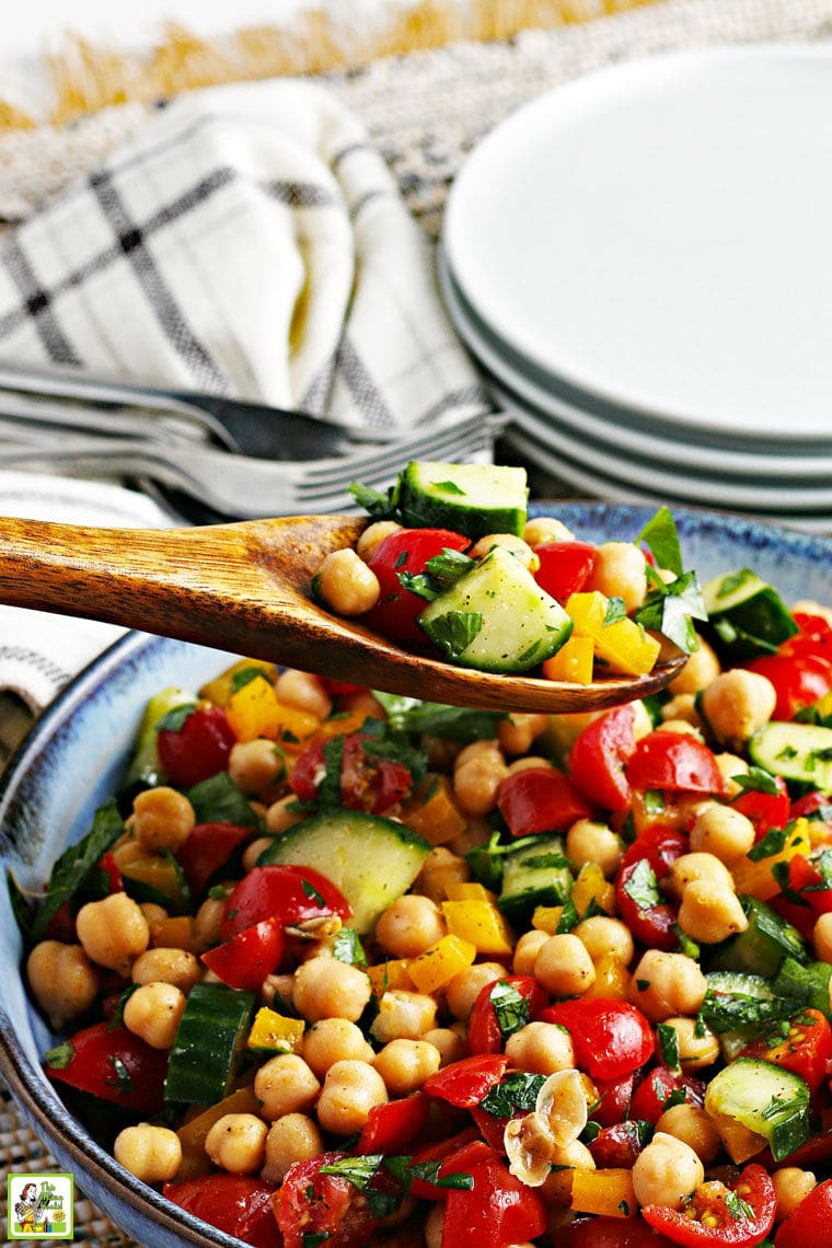 Closeup of a bowl of chickpea salad with cucumbers and tomatoes being served with a wooden spoon with white plates, forks, and white napkins in the background.