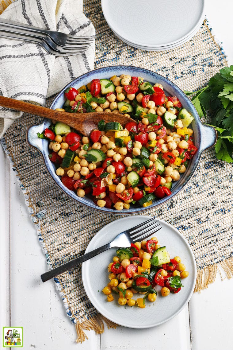 Overhead shot of a blue bowl and white plate with fork of chickpea salad with tomatoes, peppers, and cucumbers on a rustic woven gray placemat with white napkins, forks, and white plates.