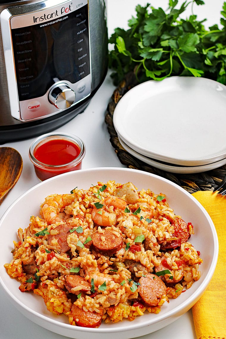 A large white bowl of jambalaya with shrimp, chicken, and sausage, a wooden spoon, a yellow napkin, ramekin of hot sauce, Instant Pot pressure cooker, a stack of white plates, and a bunch of fresh parsley.