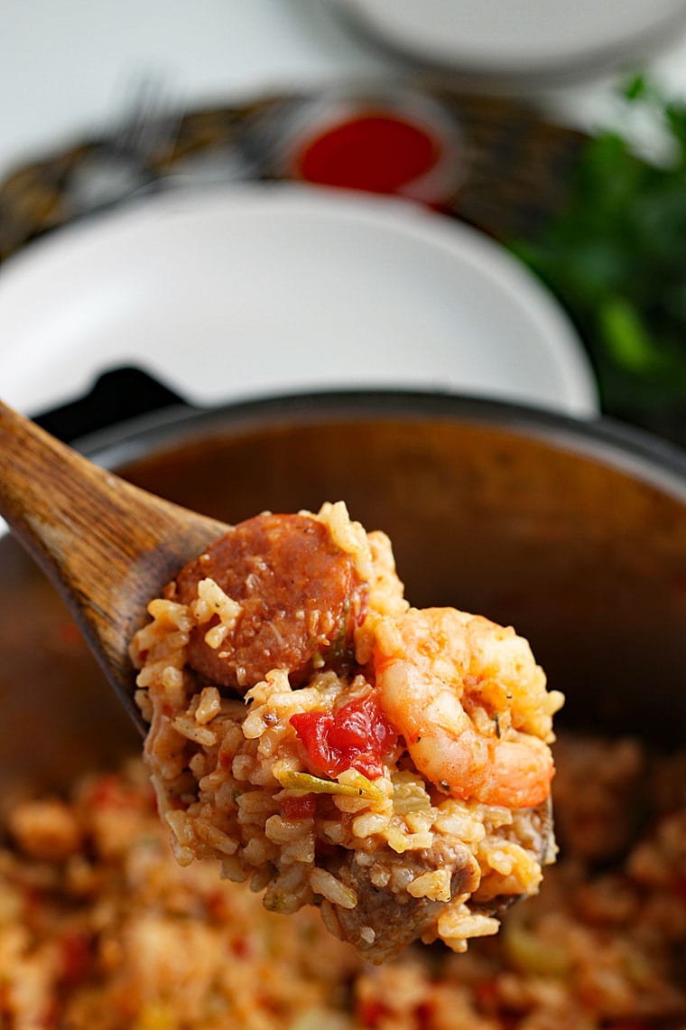 A wooden spoon filled with jambalaya with rice, shrimp, and sausage above a pressure cooker pot of jambalaya with a white plate and ramekin of hot sauce in the background.
