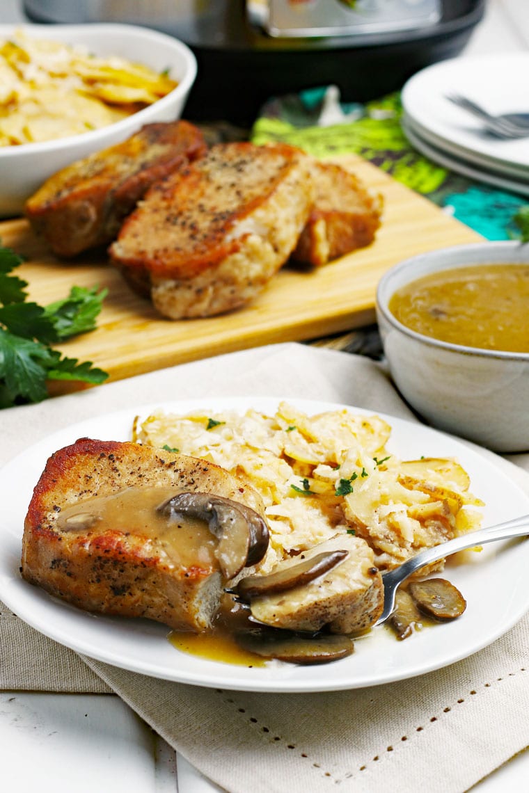 Pork chops and scallop potatoes on a white plate with fork on a linen napkin with a bowl of gravy with pork chops on a wooden cutting board in the background.