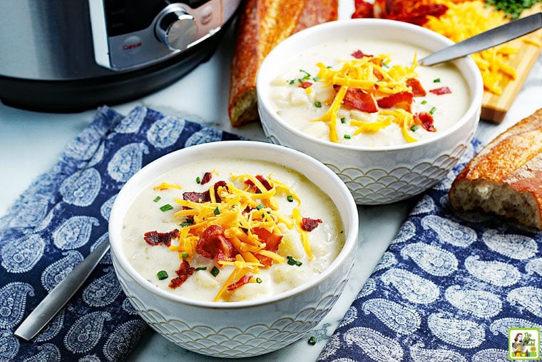 Bowls of Instant Pot potato soup with toppings of cheese and bacon bits with spoons, blue napkins, and French bread with a pressure cooker in the background.