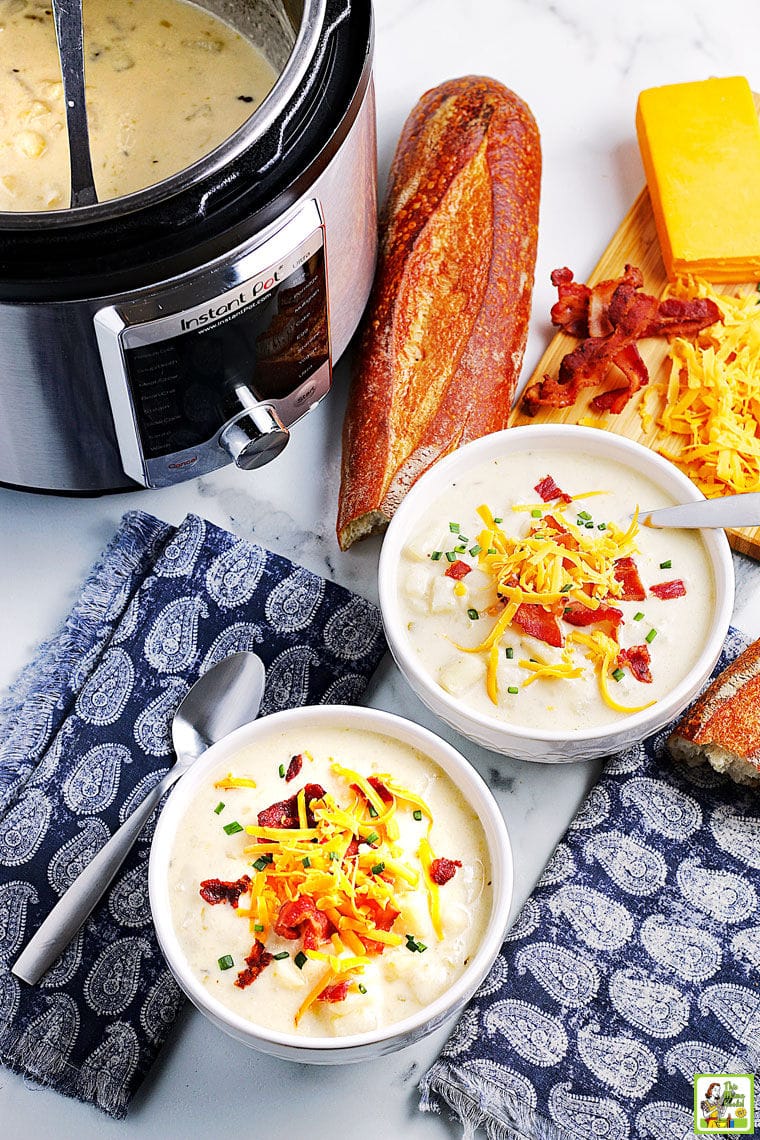 Overhead shot of bowls of potato soup topped with cheese, bacon and chives placed on blue and white napkins, next to spoons, an Instant Pot, bread, and cheddar cheese.