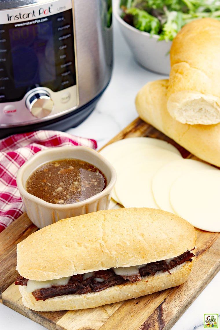 Closeup of a French dip sandwich with au jus, red and white napkin, slices of cheese and bread rolls, and an Instant Pot  and a bowl of salad in the background.