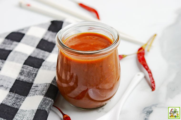 Glass jar of Homemade Hoisin Sauce with black and white napkin, red peppers, white spoon, and chopsticks.