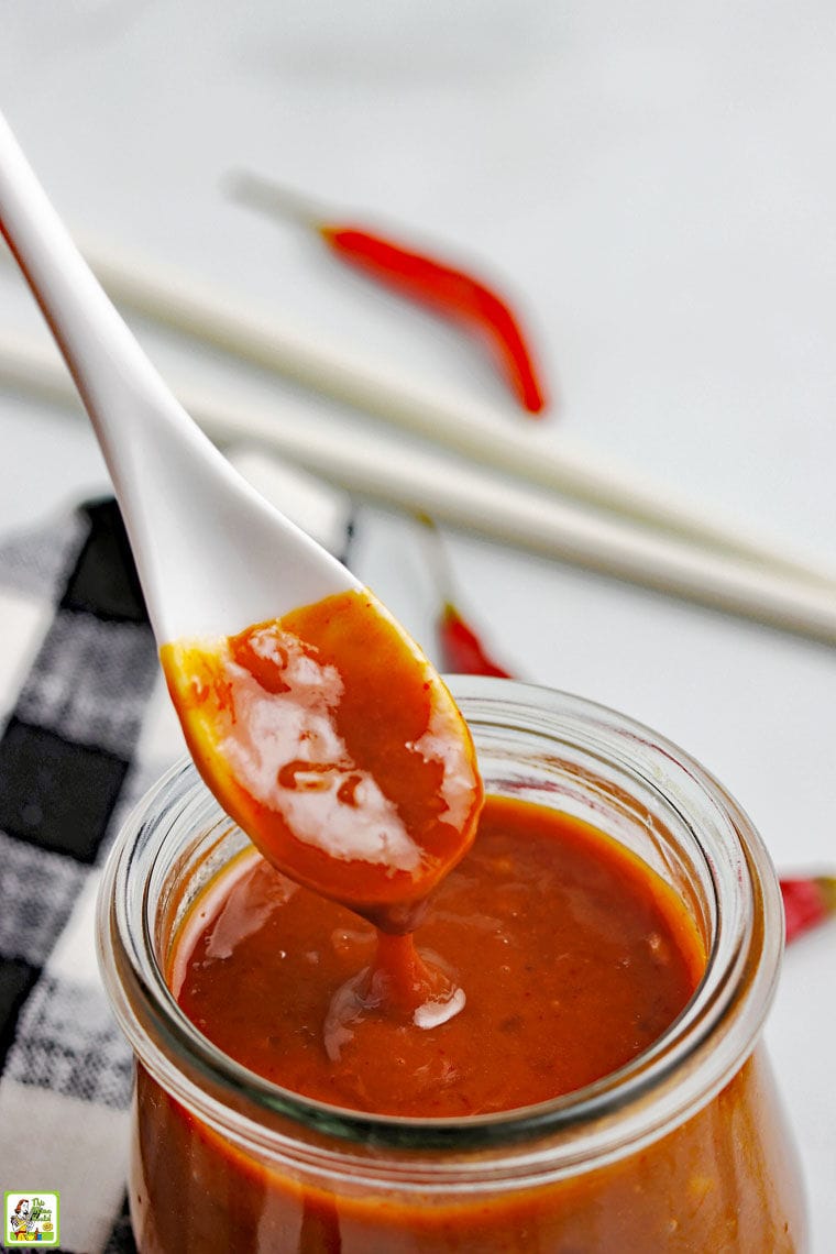 Closeup of a white spoon dipping into a glass jar of hoisin sauce with black and white napkin, red peppers, and chopsticks in the background.