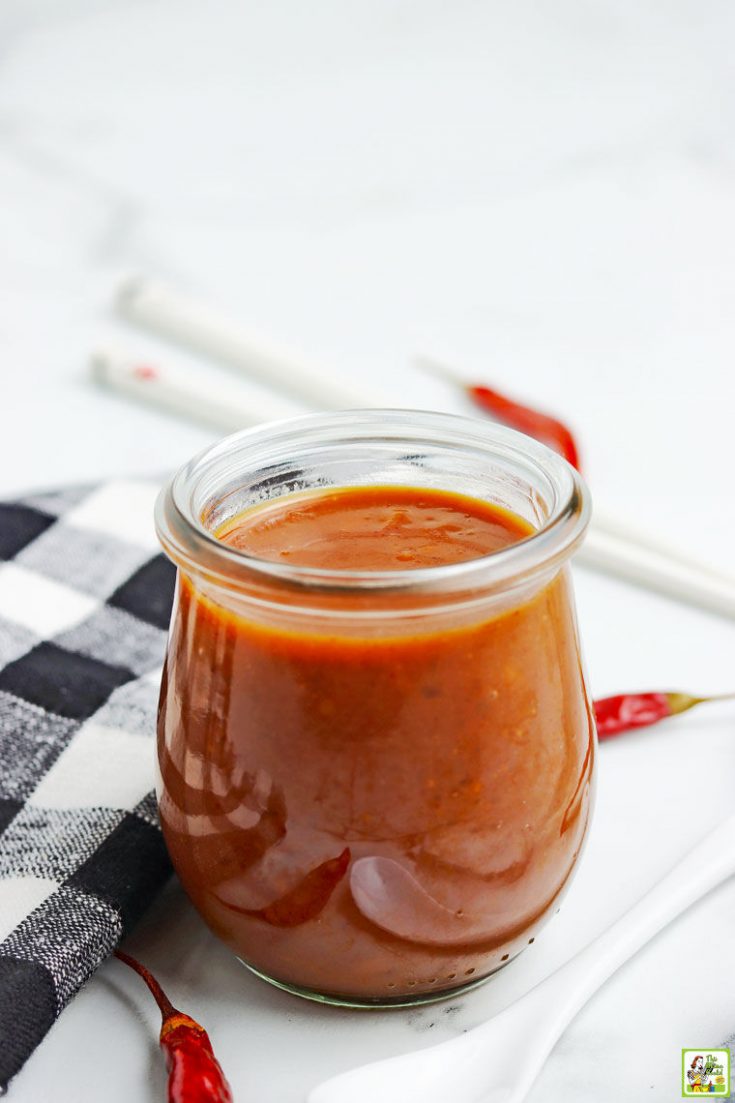 A glass jar of hoisin sauce with black and white napkin, thin red peppers, white plastic spoon, and chopsticks in the background.