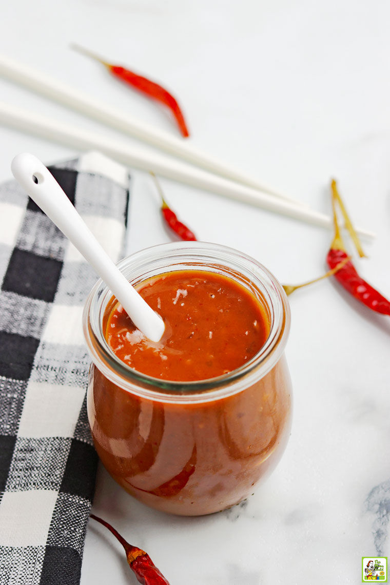 White spoon in a glass jar of homemade hoisin sauce with black and white napkin, red peppers, and chopsticks.