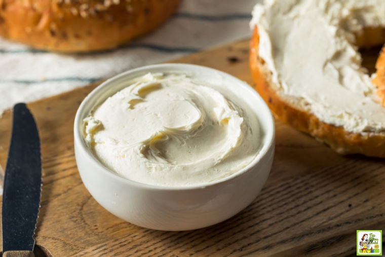 Small white bowl of cream cheese with a cream cheese covered bagel and knife on a wooden cutting board.