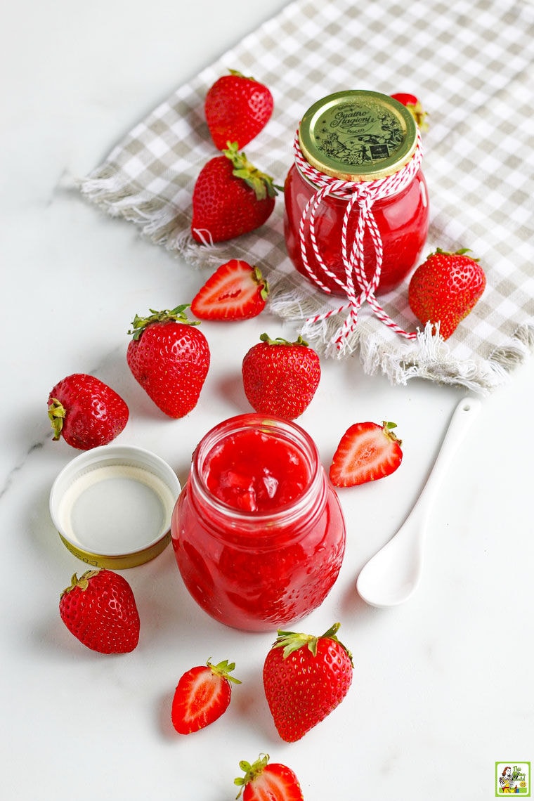 Overhead shot of glass jars of strawberry sauce with a white spoon, gray napkin, and sliced and whole strawberries.