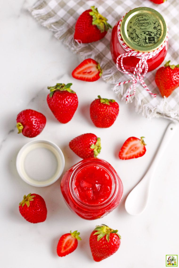 Overhead shot of glass jars of strawberry jam with a white spoon, gray napkin, and sliced and whole strawberries.