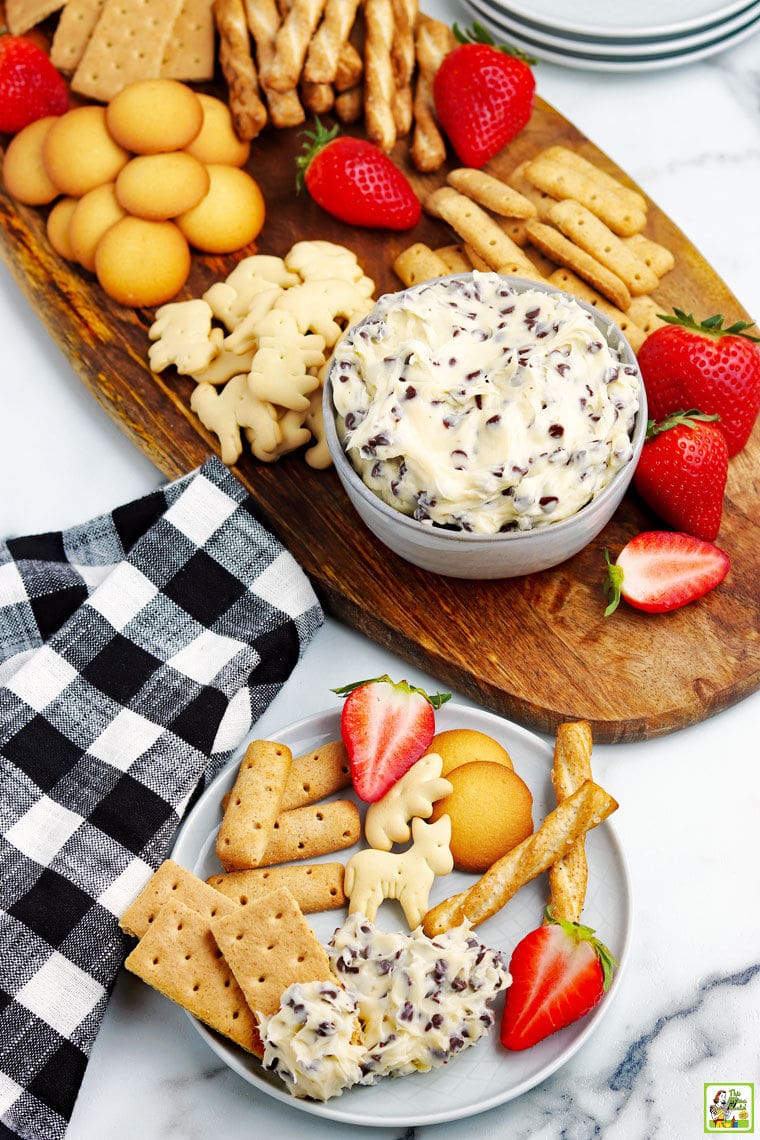 Overhead shot of a dessert board of chocolate chip cookie dough dip in a bowl and dippers of fruit, vanilla cookies, animal crackers, and cookie dough dip and dippers on a plate in the foreground.