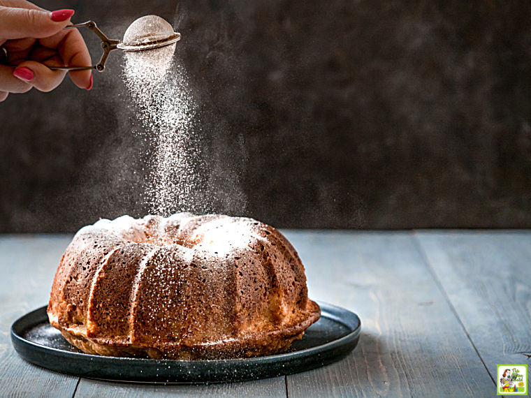 Hand sprinkling powdered sugar over a bundt cake on a dark colored plate.