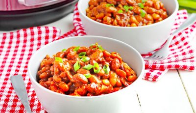 Two white bowls filled with Instant Pot Baked Beans on red and white checkered napkins with a fork and pressure cooker.