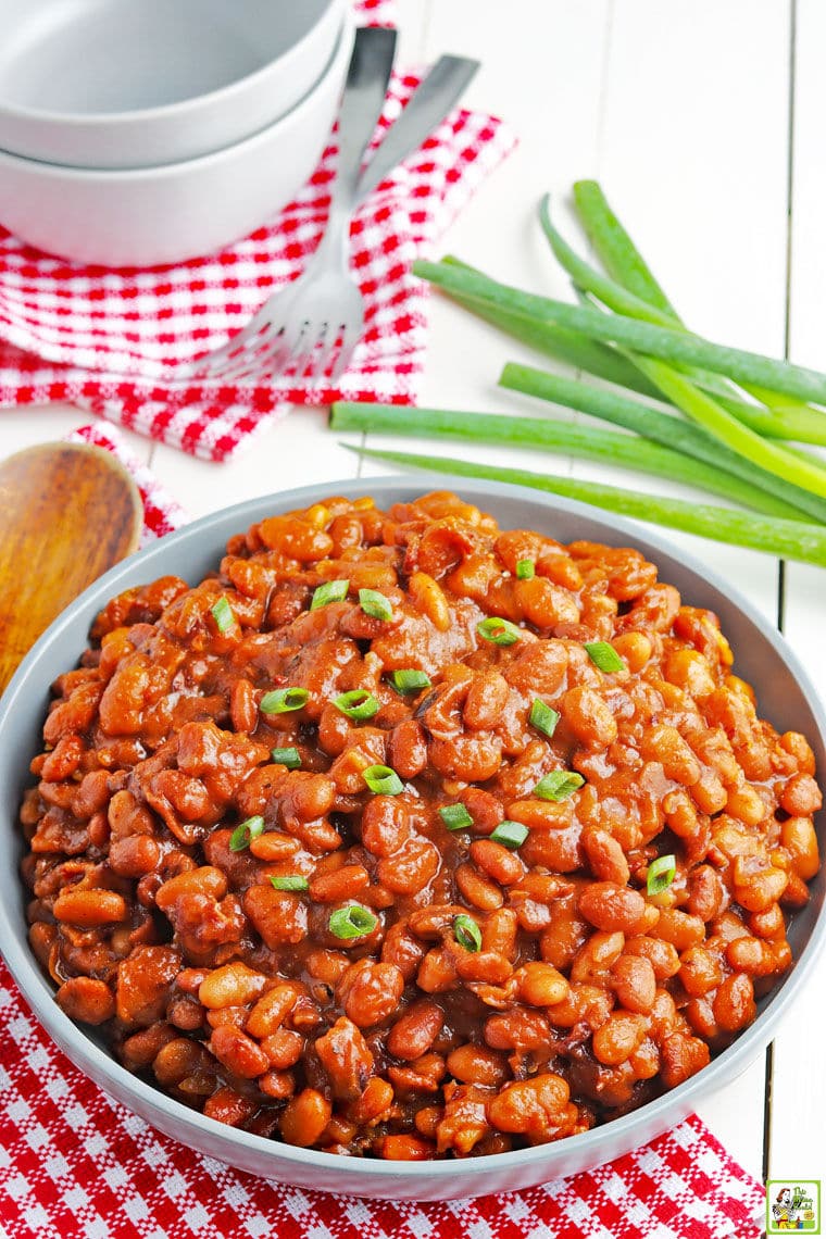 A large blue bowl of Instant Pot Baked Beans with green onions red and white checkered napkins, bowls, and forks.
