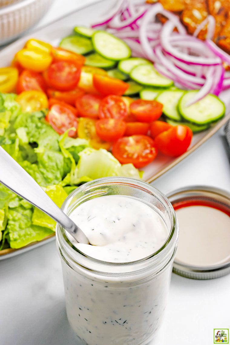 A jar of homemade ranch dressing in a mason jar with lid and spoon and a platter of salad fixings including lettuce, sliced tomatoes, cucumbers, and onions.