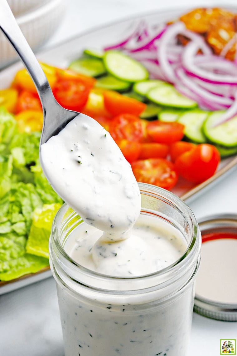 A spoon dipped into a jar of homemade ranch dressing in a mason jar with with a platter of salad fixings including lettuce, sliced tomatoes, cucumbers, and onions in the background.