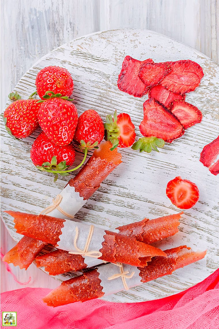Piles of strawberry fruit leather roll ups, strawberries, and strawberry chips on a white wooden cutting board.