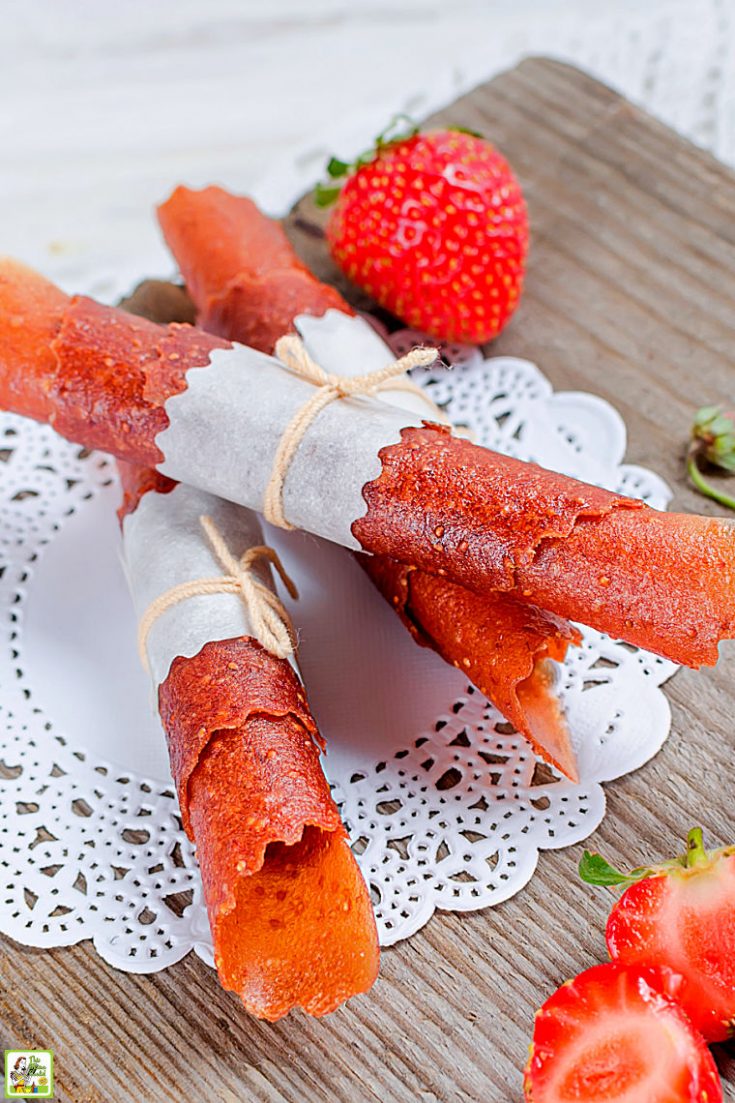 Strawberry fruit leather roll ups wrapped in parchment and string on a lace doily on a wooden cutting board with strawberries.
