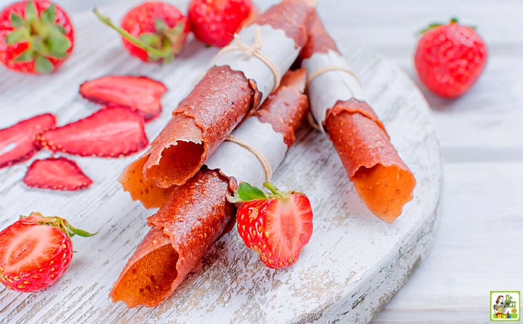 Strawberries, strawberry fruit leather roll ups wrapped in white parchment paper and string, and dried strawberry chips on a white wooden board.