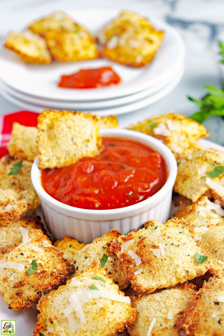 A small bowl of marinara sauce with toasted air fryer ravioli with a plate of toated ravioli in the background.