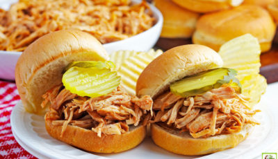 Two Instant Pot Pulled BBQ Chicken Sandwiches with pickles and potato chips on a white plate with bowl of shredded chicken and buns in the background.