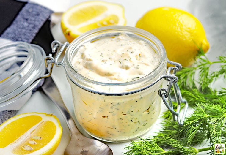 A glass jar of Tartar Sauce with sliced lemons, fresh dill, a spoon, and black and white napkin.