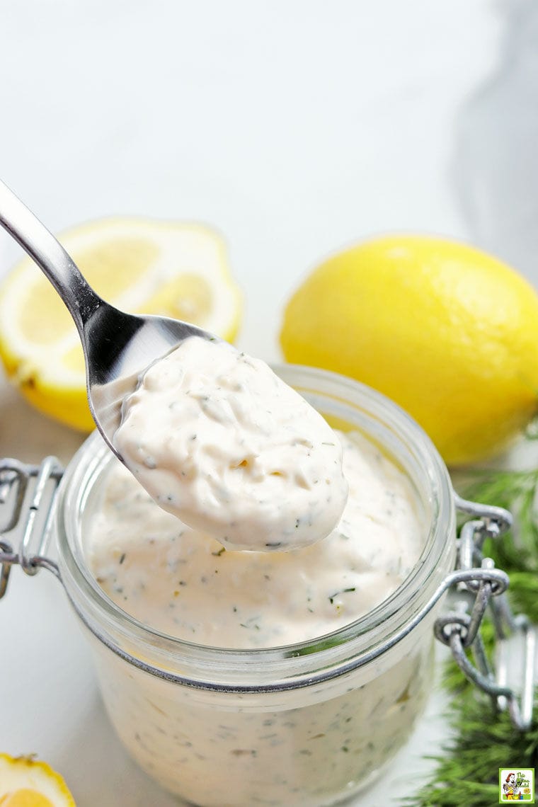 A spoon dipping in a glass jar of tartar sauce with lemons and fresh dill.