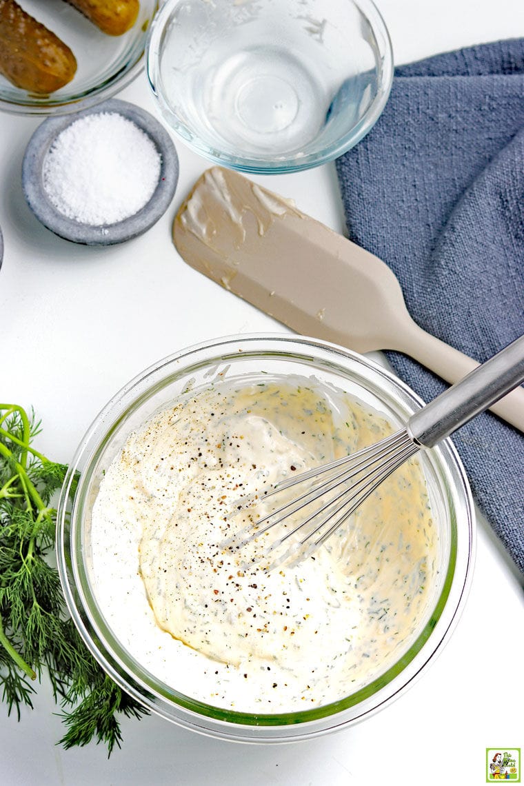 Overhead view of mixing bowl of tartar sauce with whisk, spatula, fresh dill, a ramekin of salt, a small glass bowl, and a blue kitchen towel.