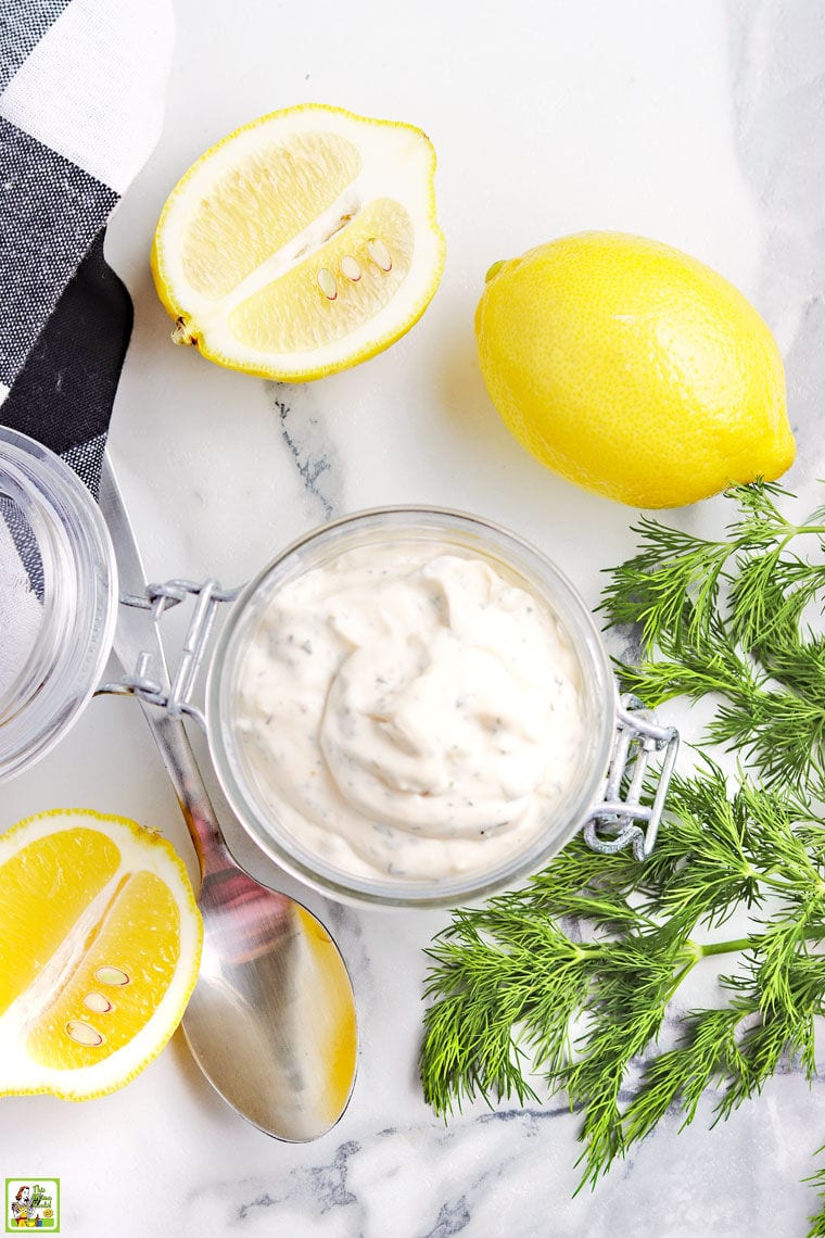 Overhead view of a glass jar filled with homemade tartar sauce, fresh dill, sliced and whole lemons, and a black and white cloth napkin.