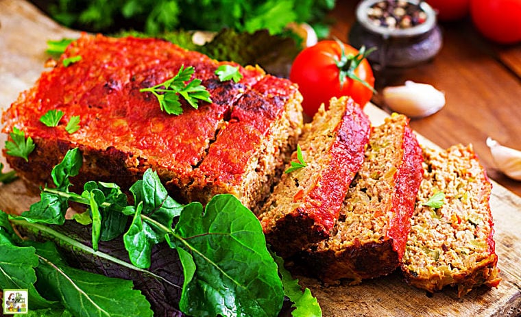 Sliced loaf of meat loaf covered in tomato sauce on a wooden cutting board with fresh herbs.