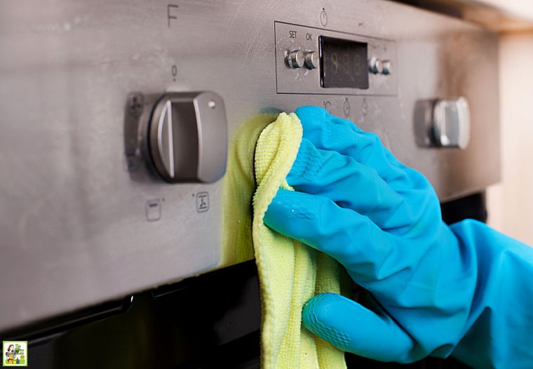 A hand wearing a blue rubber glove wiping down oven dials with a cleaning rag.