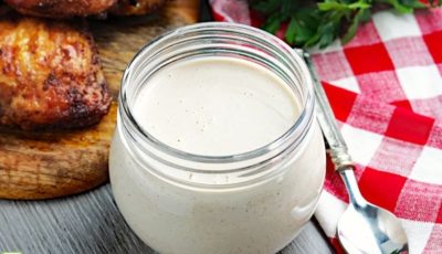 A small jar of Alabama white BBQ sauce with spoon, gingham napkin, and barbecue chicken.