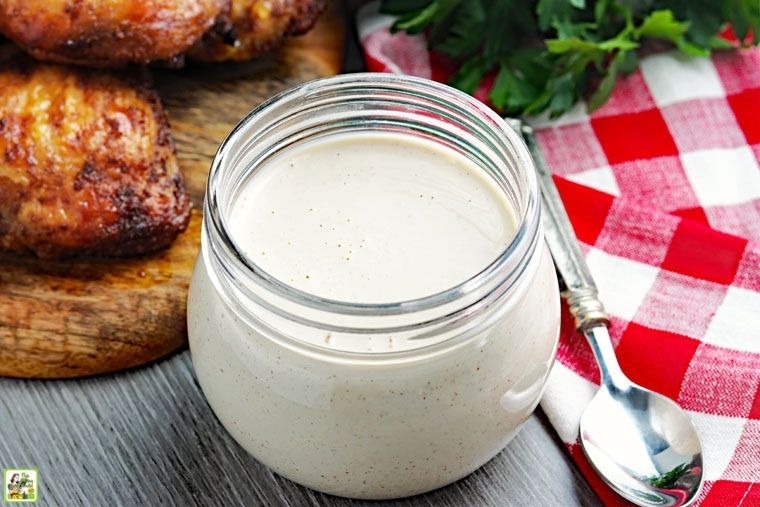 A small jar of Alabama white BBQ sauce with spoon, gingham napkin, and barbecue chicken.