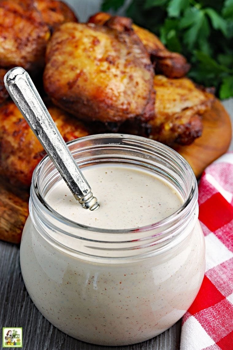 A glass jar of Alabama white sauce with spoon and red and white gingham napkin and barbecue chicken.