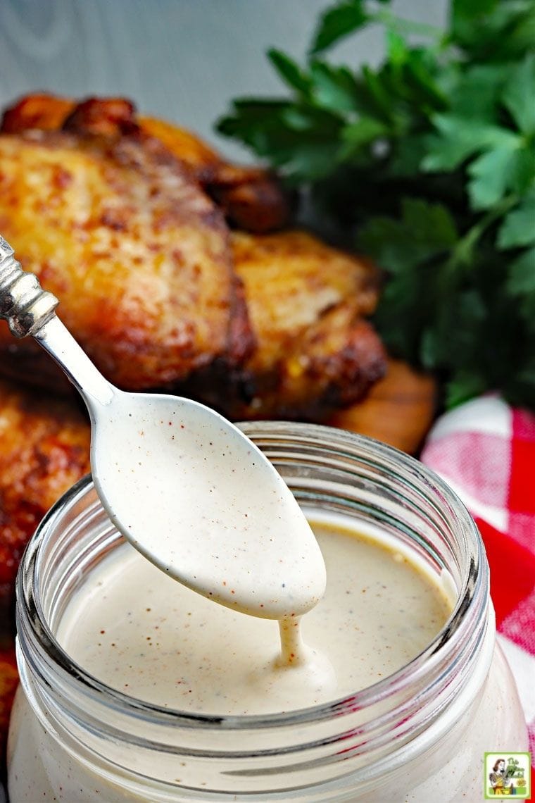 Spoon dipped into a jar of Alabama white BBQ sauce a pile of barbecue chicken in the background.