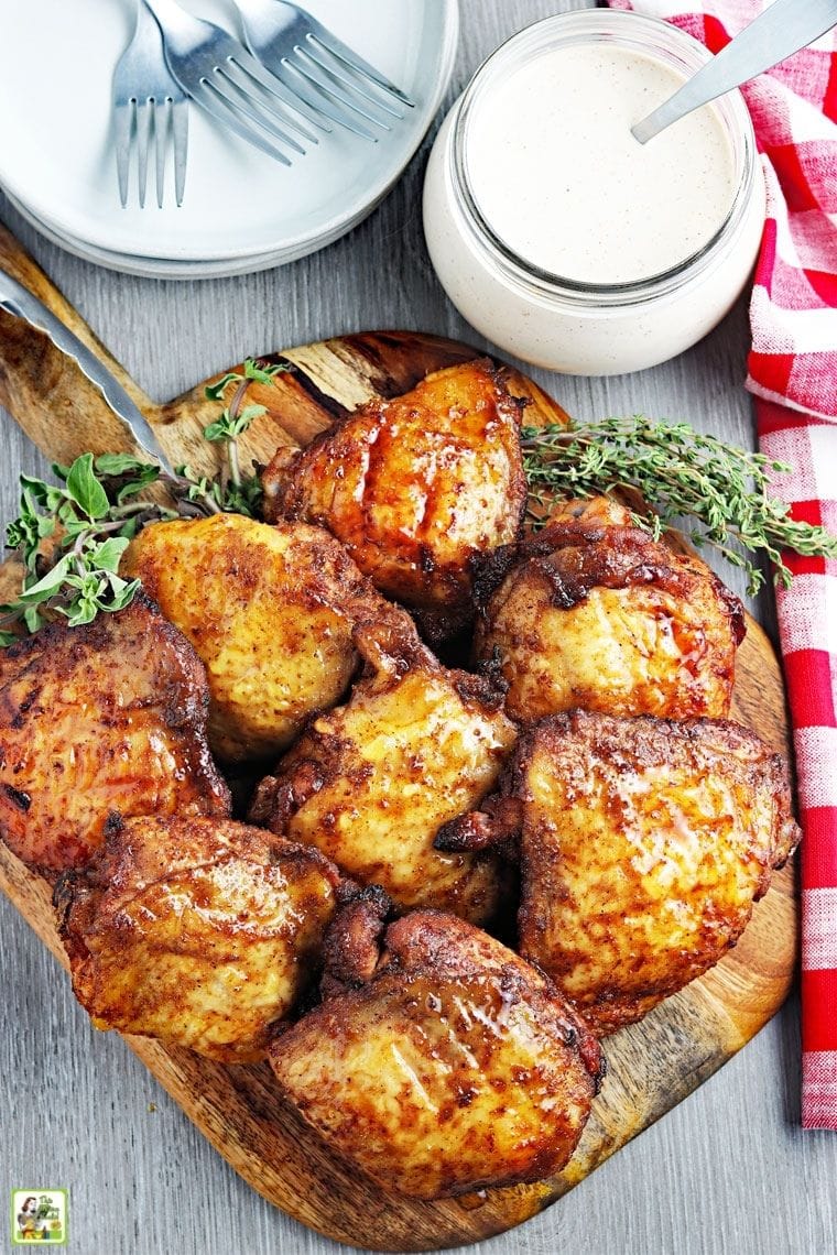 Κάτοψη των μαγειρεμένων καπνιστών μηρών κοτόπουλου σε μια ξύλινη σανίδα κοπής με ένα βάζο με σάλτσα Αλαμπάμα, πιάτα, σκεύη και κόκκινες και άσπρες υφασμάτινες χαρτοπετσέτες.