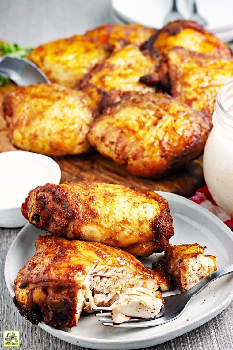 Ένα πιάτο με καπνιστούς μηρούς κοτόπουλου με περισσότερο κοτόπουλο στο βάθος και βάζα με λευκή σάλτσα μπάρμπεκιου.