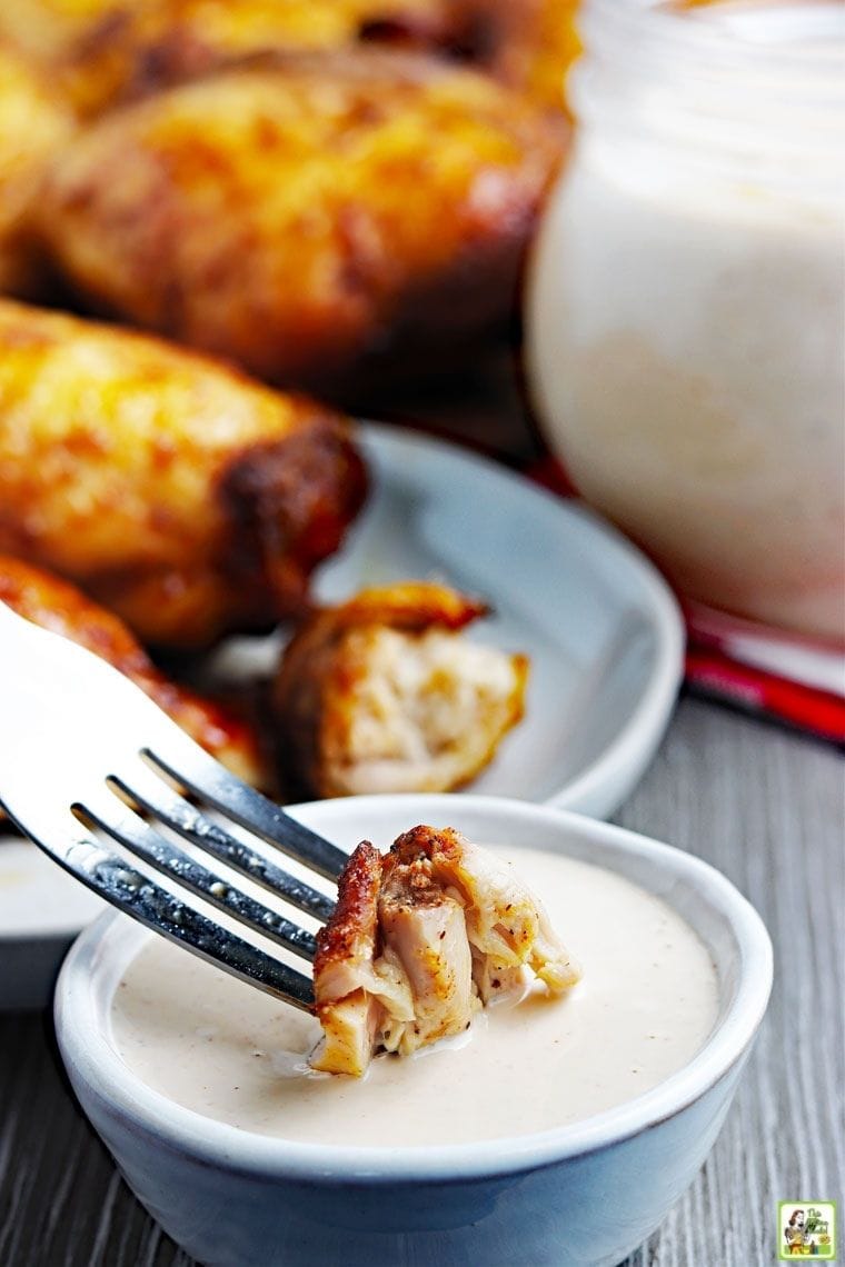 Ένα κομμάτι μαγειρεμένο κοτόπουλο σε ένα πιρούνι που βυθίζεται σε σάλτσα μπάρμπεκιου Αλαμπάμα με κομμάτια μαγειρεμένων καπνιστών μηρών κοτόπουλου στο βάθος.